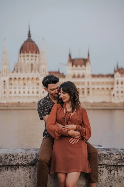 Budapest engagement photoshoot: An embrace from behind opposite Parliament The couple embracing from behind and looking down with gentle smiles, with Parliament softly blurred across the river during their Budapest engagement photoshoot.