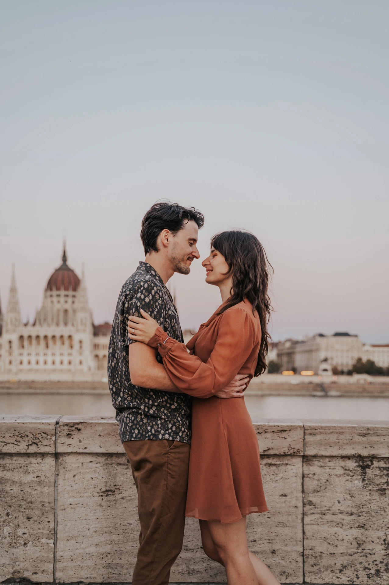 Budapest engagement photoshoot: An embrace opposite Parliament The couple embracing beside the stone river wall, with the Hungarian Parliament’s ornate façade behind them across the Danube during their Budapest engagement photoshoot.