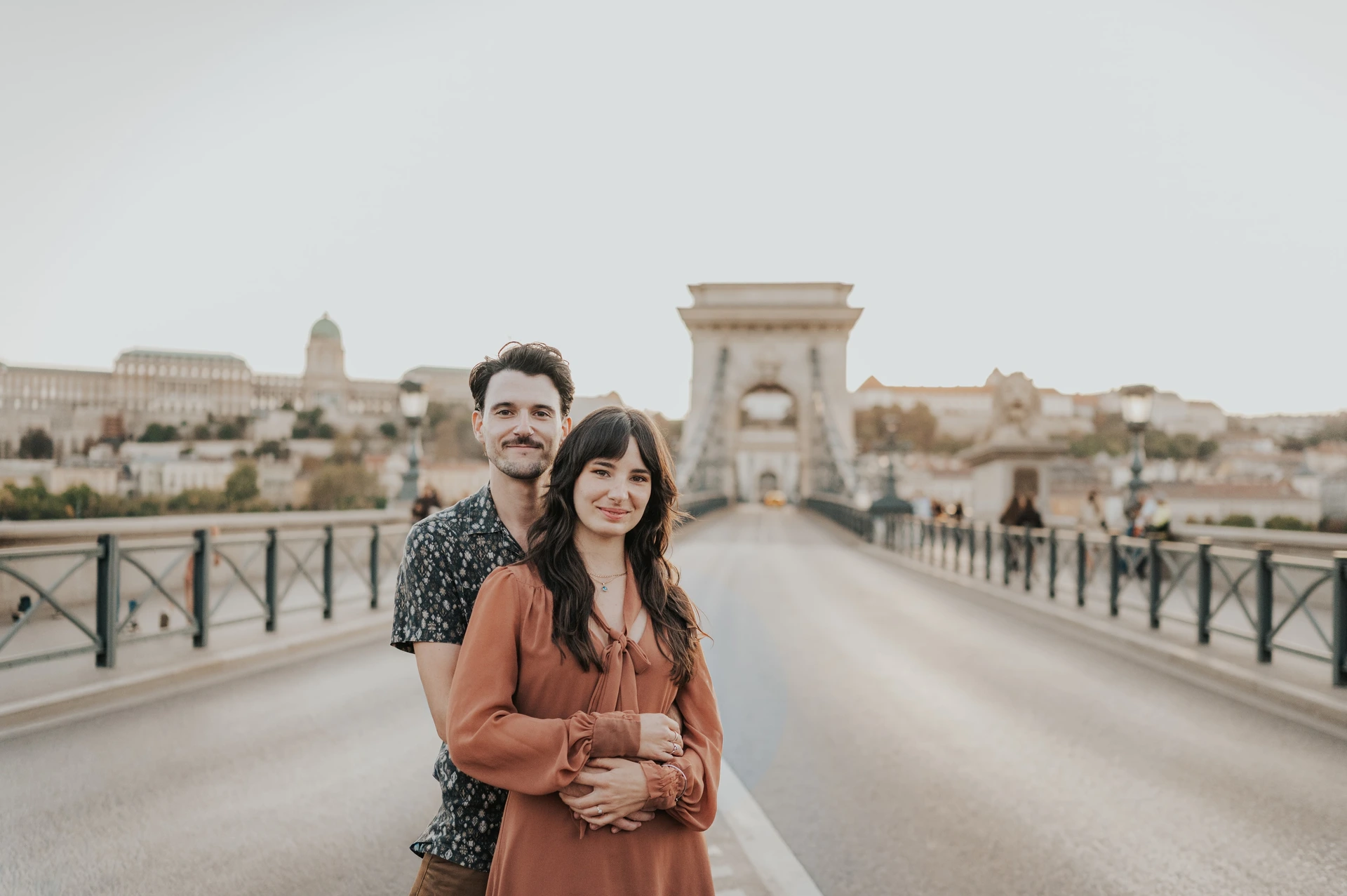 Budapest engagement photoshoot: An embrace with Chain Bridge stretching behind A landscape portrait of the couple embracing on Chain Bridge, looking towards the camera with the bridge tower in the background during their Budapest engagement photoshoot.