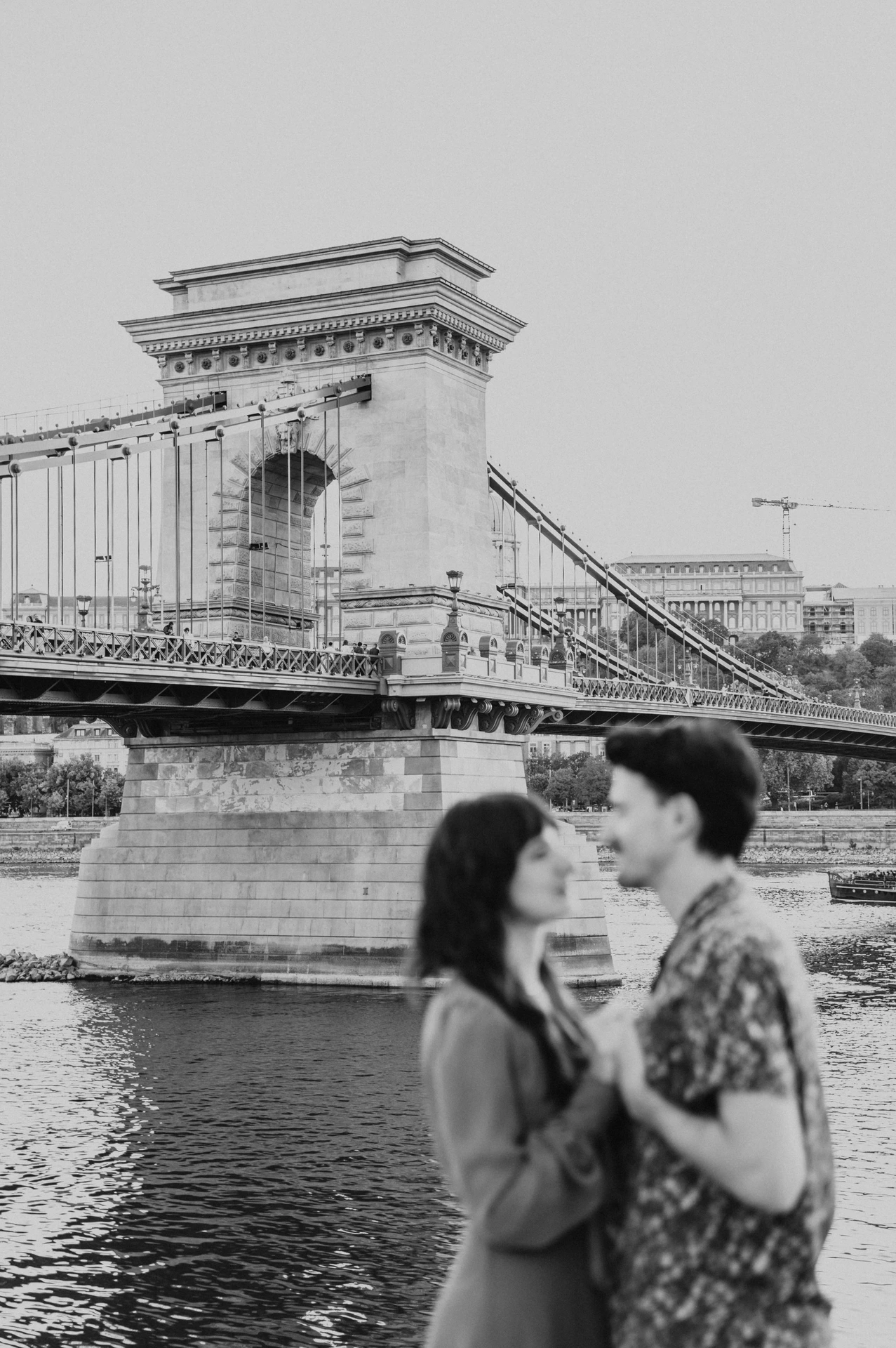 Budapest engagement photoshoot: Black-and-white closeness beside the Danube Black-and-white photograph of the couple holding hands beside the Danube, with Chain Bridge behind them during their Budapest engagement photoshoot.