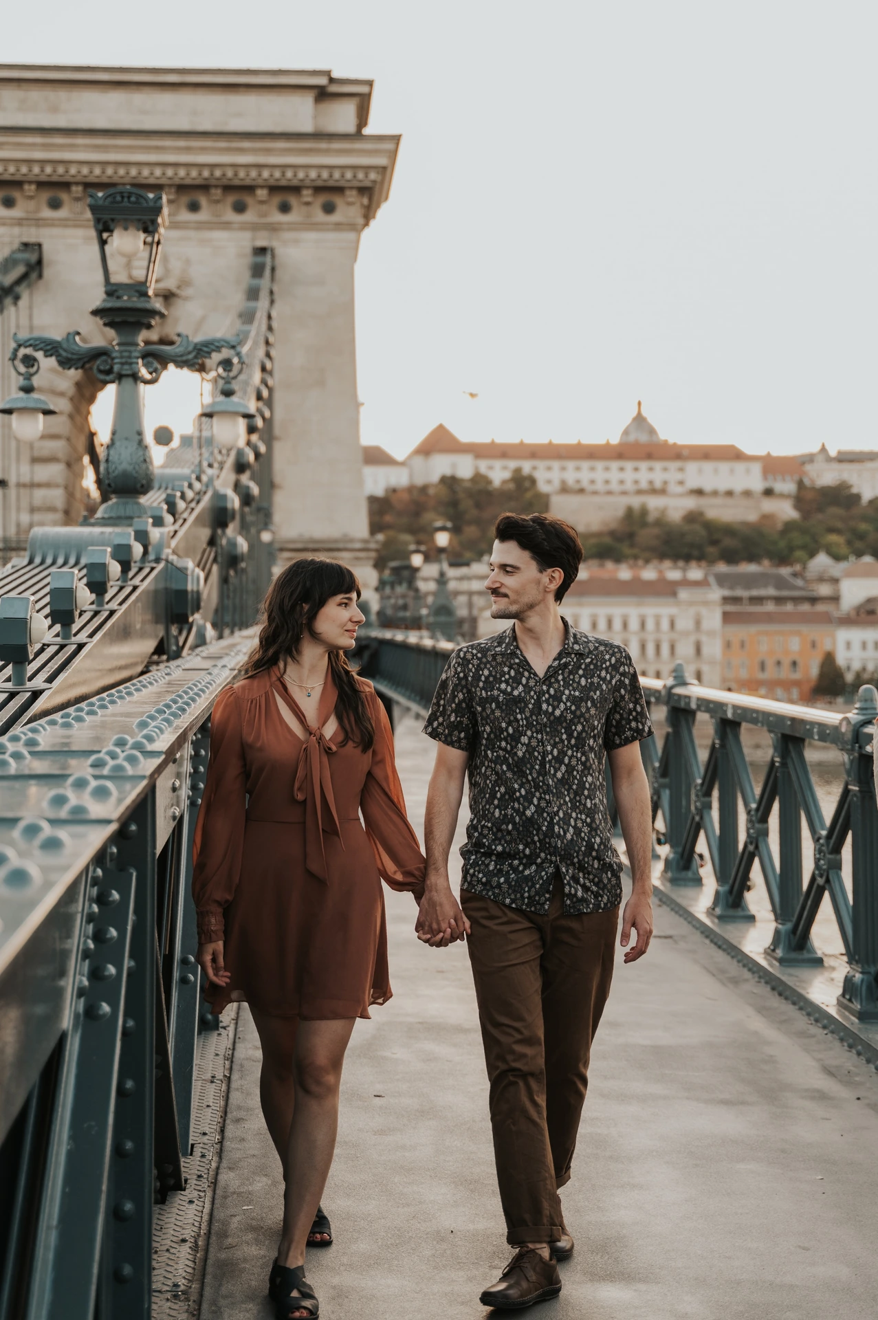 The couple walking together past the decorative lamps on Chain Bridge, holding hands and looking towards each other during their Budapest engagement photoshoot.