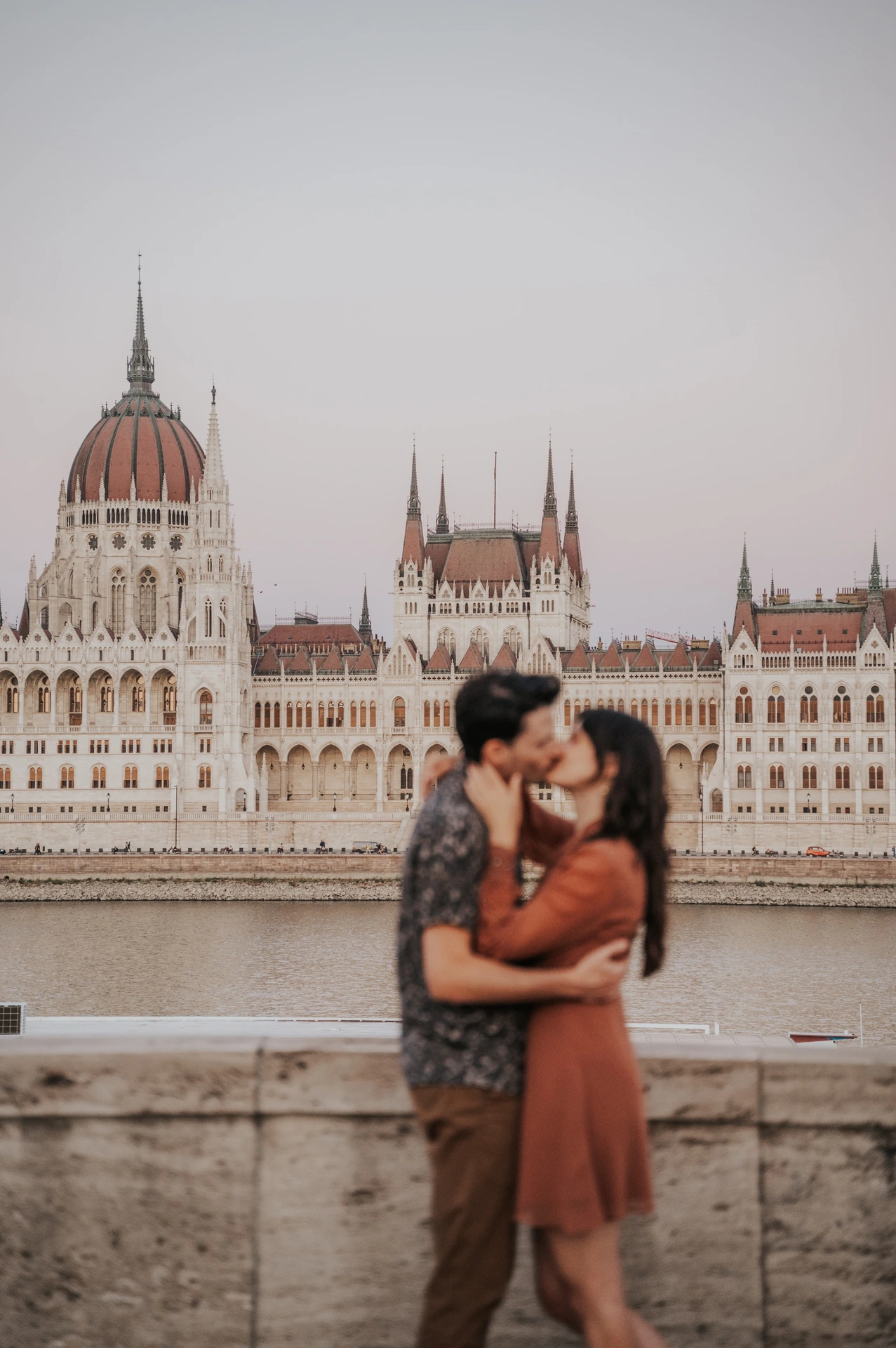 Budapest engagement photoshoot: Holding each other opposite Parliament The couple embracing and sharing a kiss by the Danube, with the Hungarian Parliament in the background during their Budapest engagement photoshoot.