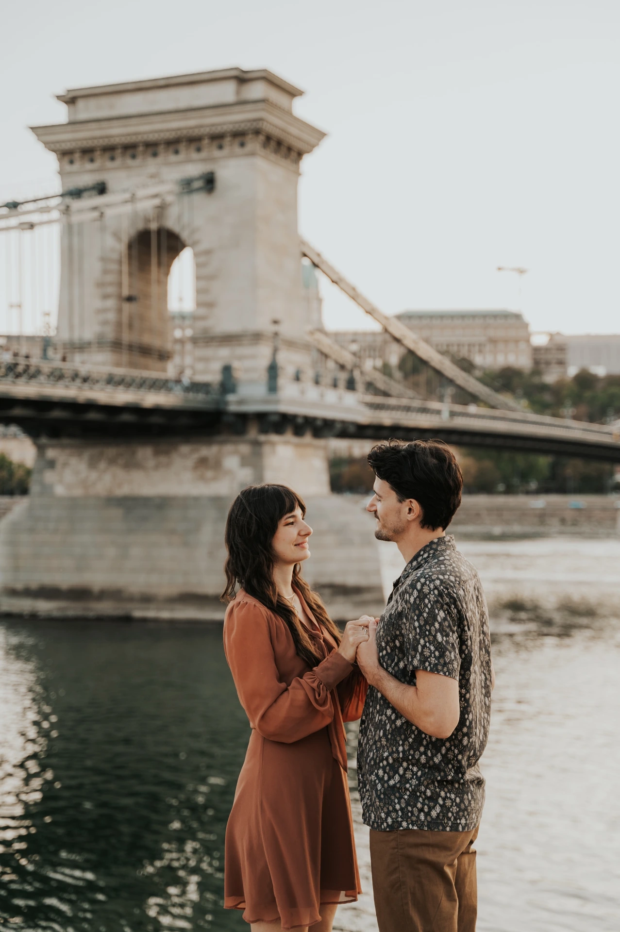 The couple facing each other and holding hands beside the Danube, with Chain Bridge’s stone tower and suspension chains behind them during their Budapest engagement photoshoot.