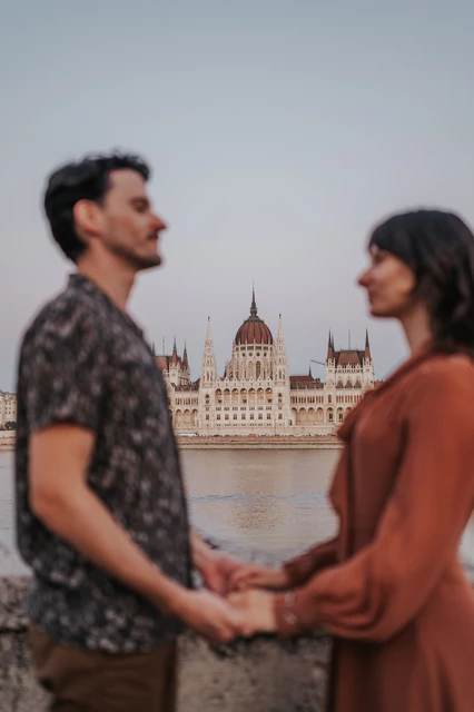 Budapest engagement photoshoot: Holding hands beside the Parliament river view The couple facing one another and holding hands beside the Danube, with Parliament’s dome in the background during their Budapest engagement photoshoot.