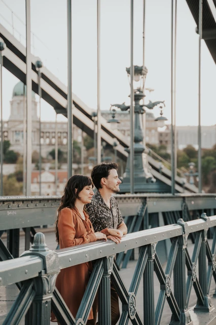 Budapest engagement photoshoot: Leaning together on Chain Bridge’s railing The couple leaning side by side on Chain Bridge’s railing and looking across the river, framed by its suspension chains during their Budapest engagement photoshoot.
