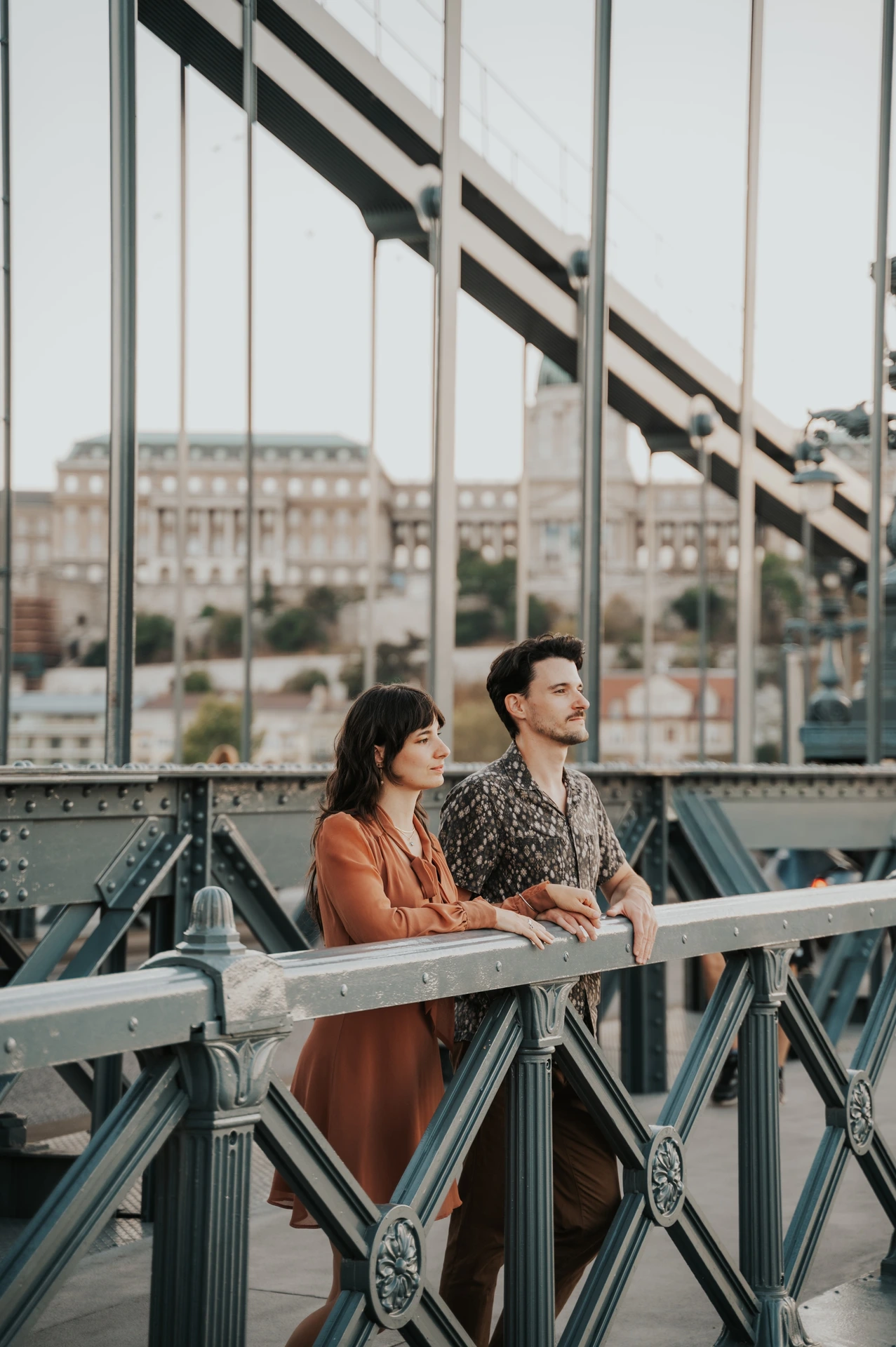 Budapest engagement photoshoot: Looking out from Chain Bridge The couple standing close at the bridge railing and looking towards the river, with suspension chains crossing the frame during their Budapest engagement photoshoot.
