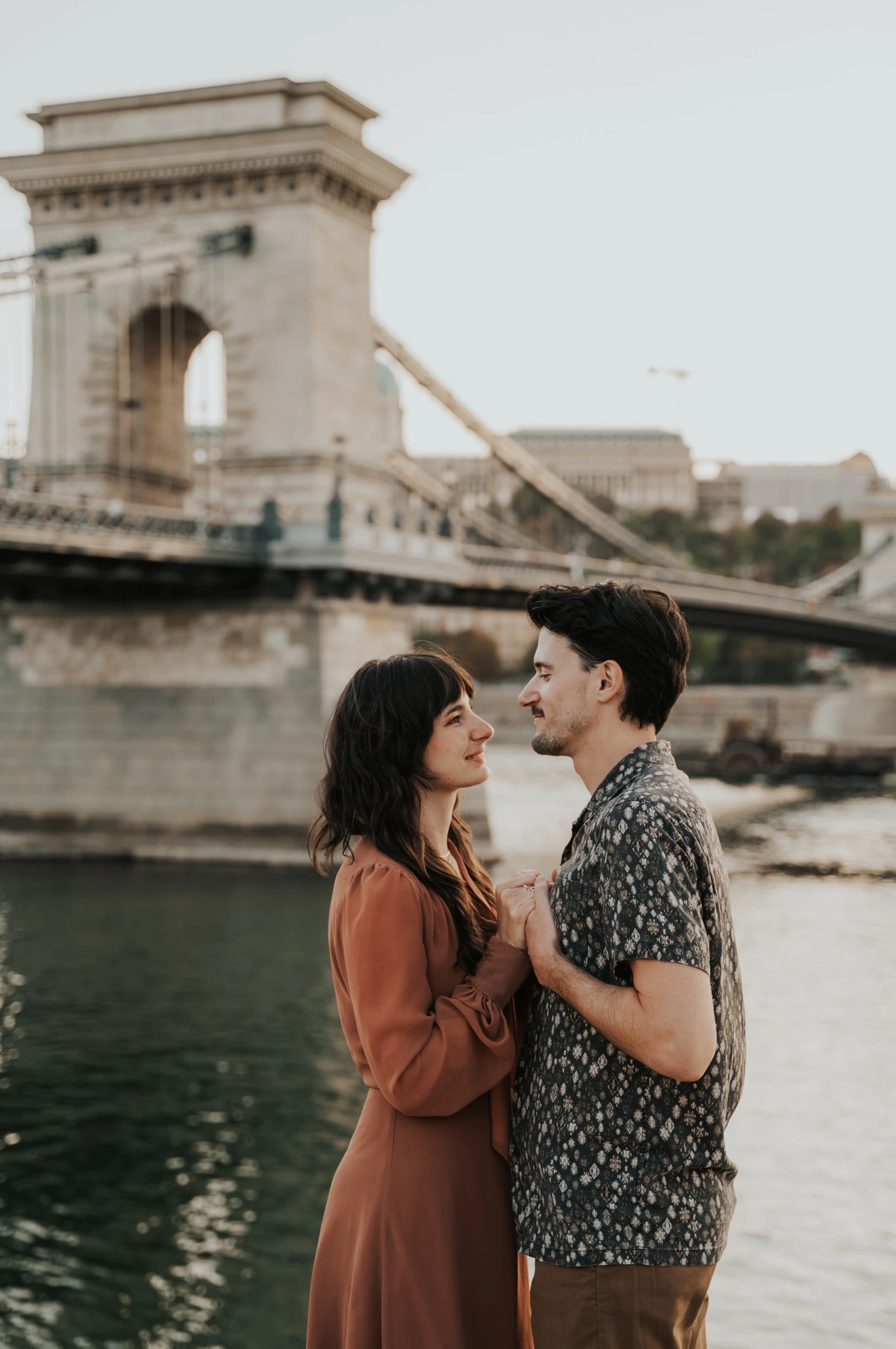 Budapest engagement photoshoot: Looking towards each other by Chain Bridge The couple sharing a look while holding hands on the riverside, with Chain Bridge’s stone tower reflected in the water behind them during their Budapest engagement photoshoot.