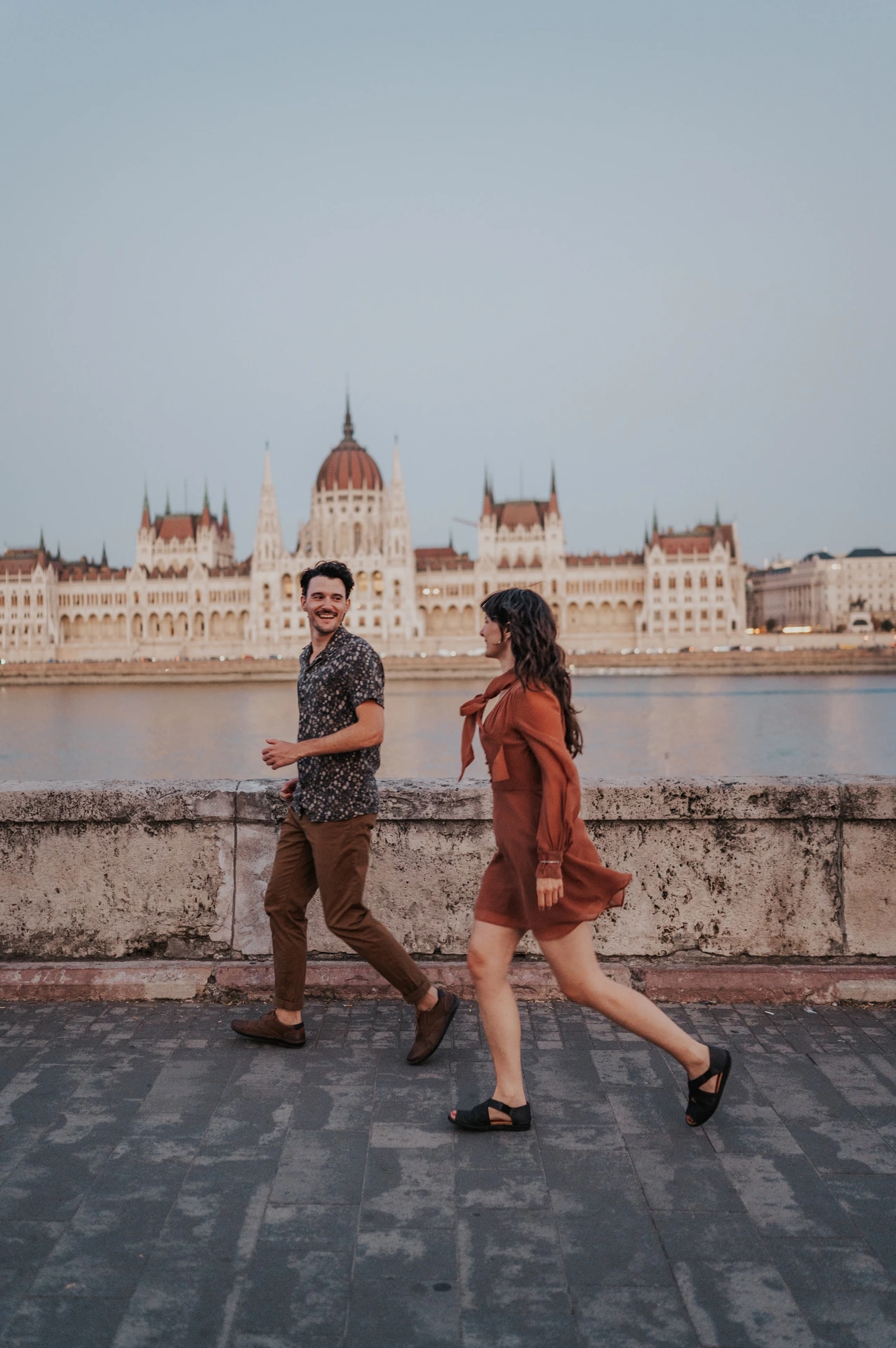 Budapest engagement photoshoot: Looking towards each other on their riverside walk The couple smiling at one another while walking beside the stone river wall, with the Hungarian Parliament behind them during their Budapest engagement photoshoot.