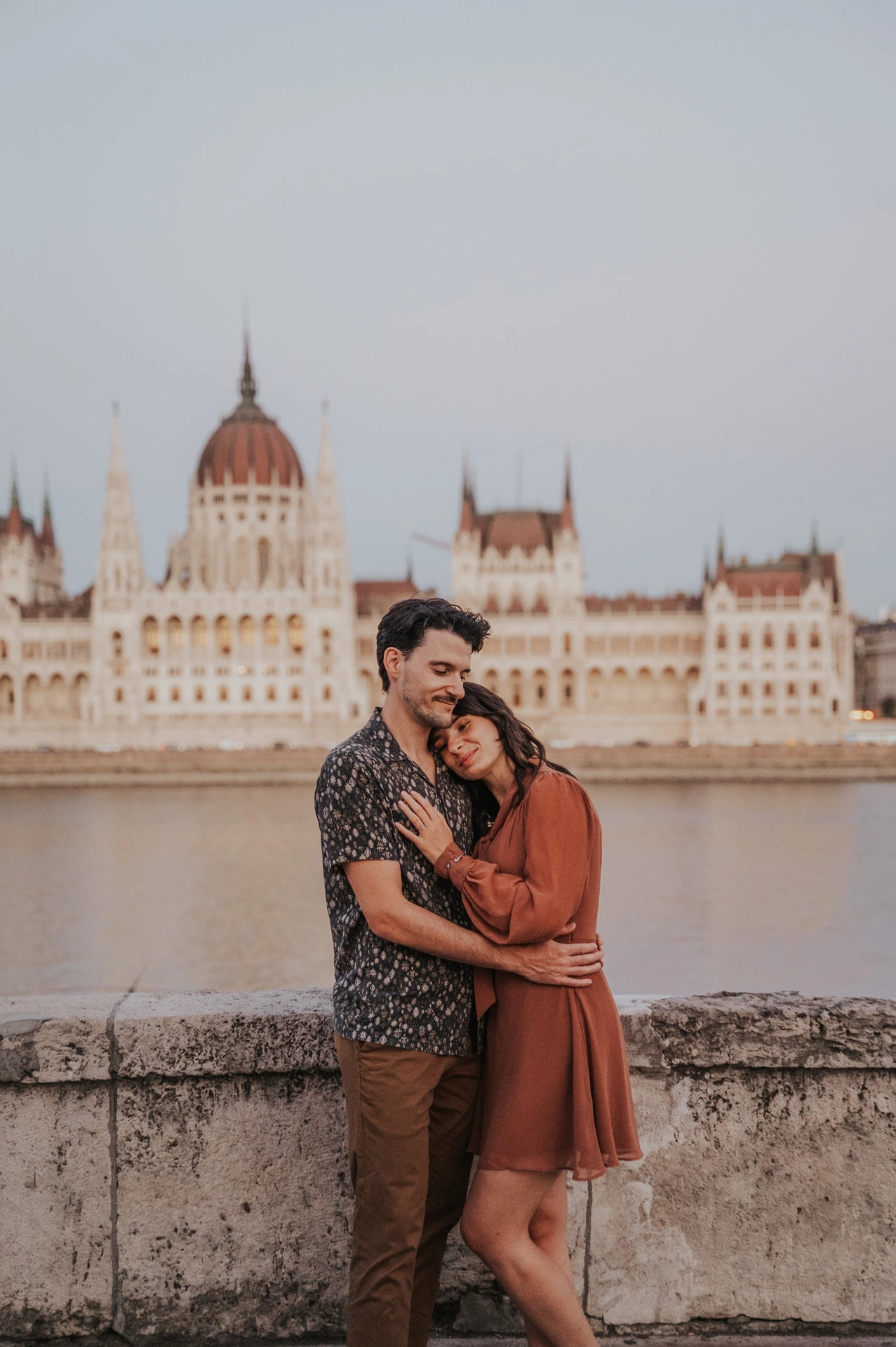 Budapest engagement photoshoot: Resting close with Parliament behind them The couple cuddling beside the river wall, with one partner resting against the other and Parliament across the Danube during their Budapest engagement photoshoot.