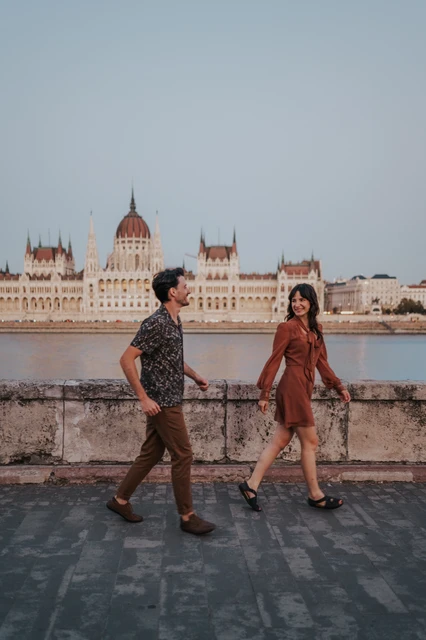 Budapest engagement photoshoot: Shared smiles along the Parliament river view The couple looking towards one another as they walk hand in hand along the Danube, with Parliament in the background during their Budapest engagement photoshoot.