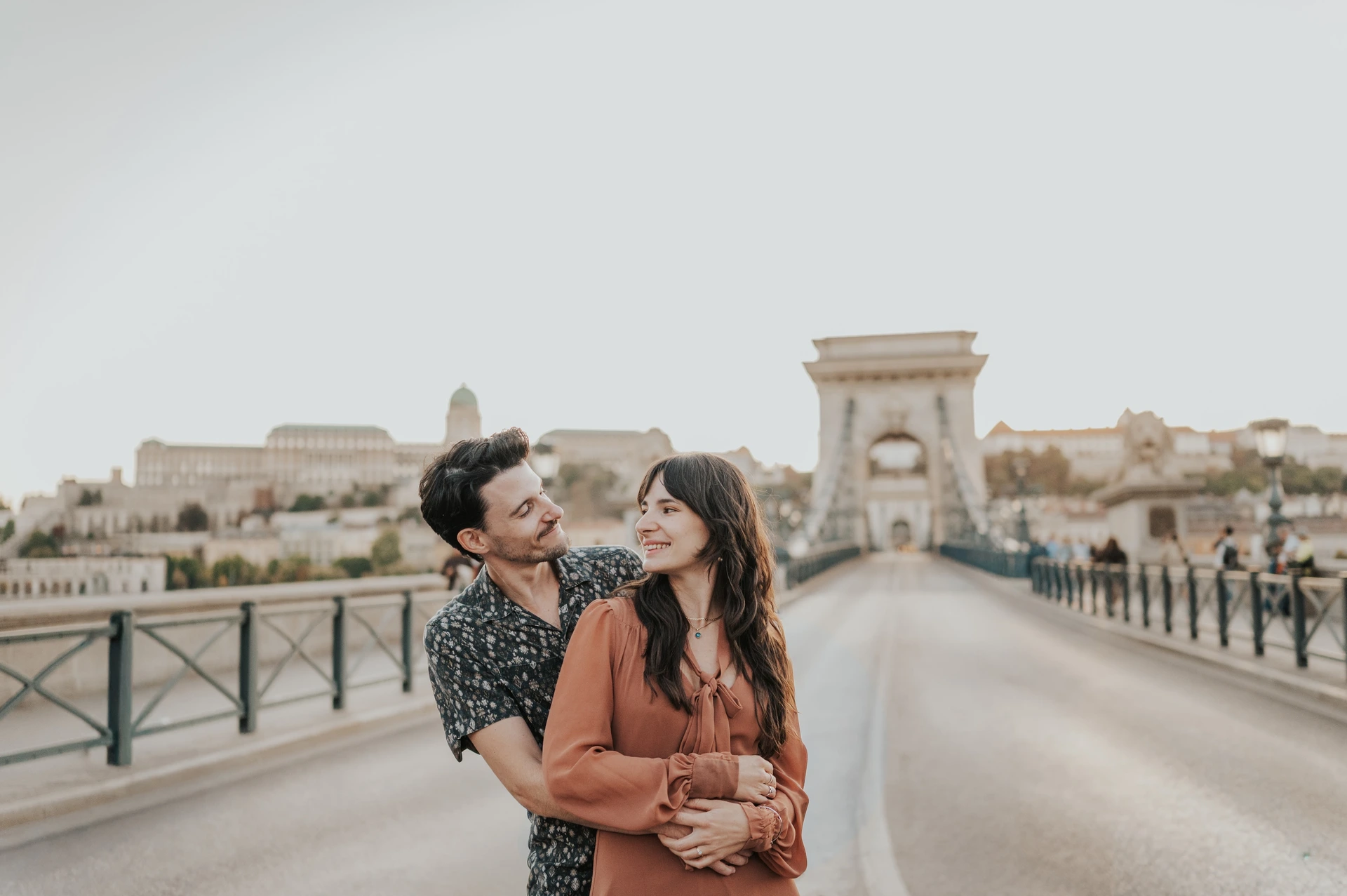 A landscape photograph of the couple smiling at one another in an embrace, with Chain Bridge’s tower and railings stretching behind them during their Budapest engagement photoshoot.