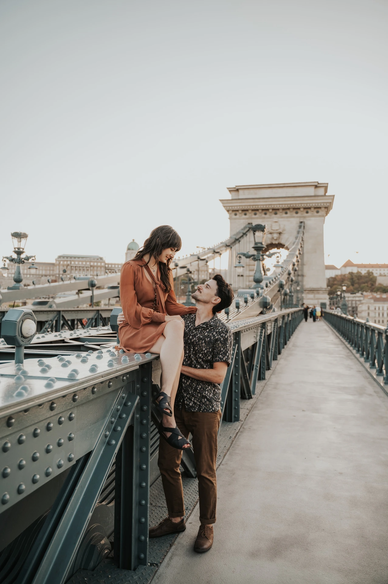 Budapest engagement photoshoot: Sharing a smile beside the bridge tower The couple smiling together beside the bridge railing, with Chain Bridge’s stone tower rising behind them during their Budapest engagement photoshoot.
