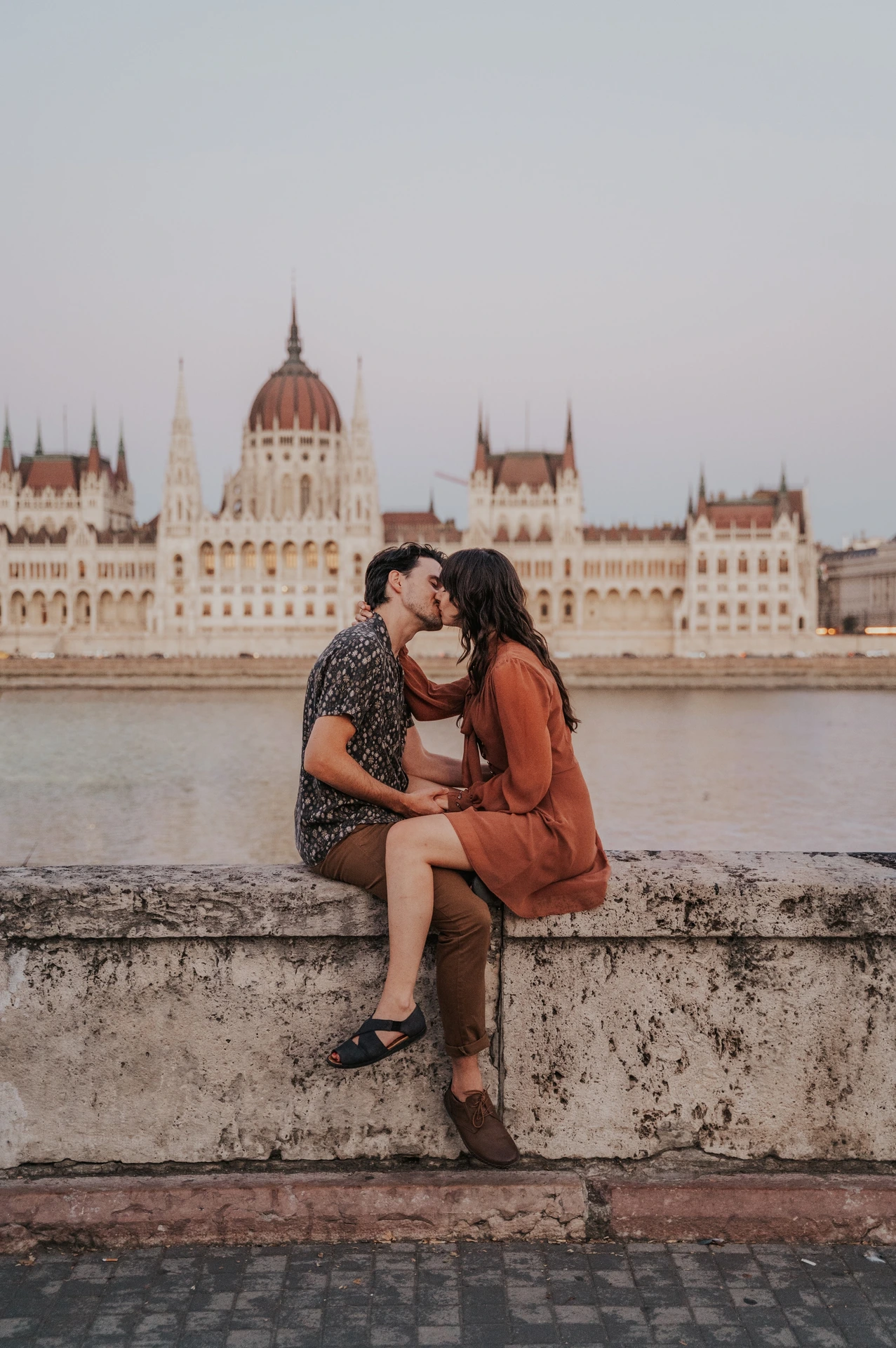 Budapest engagement photoshoot: Sitting close on the riverside wall The couple sitting closely together on the stone river wall, with the Hungarian Parliament across the Danube behind them during their Budapest engagement photoshoot.