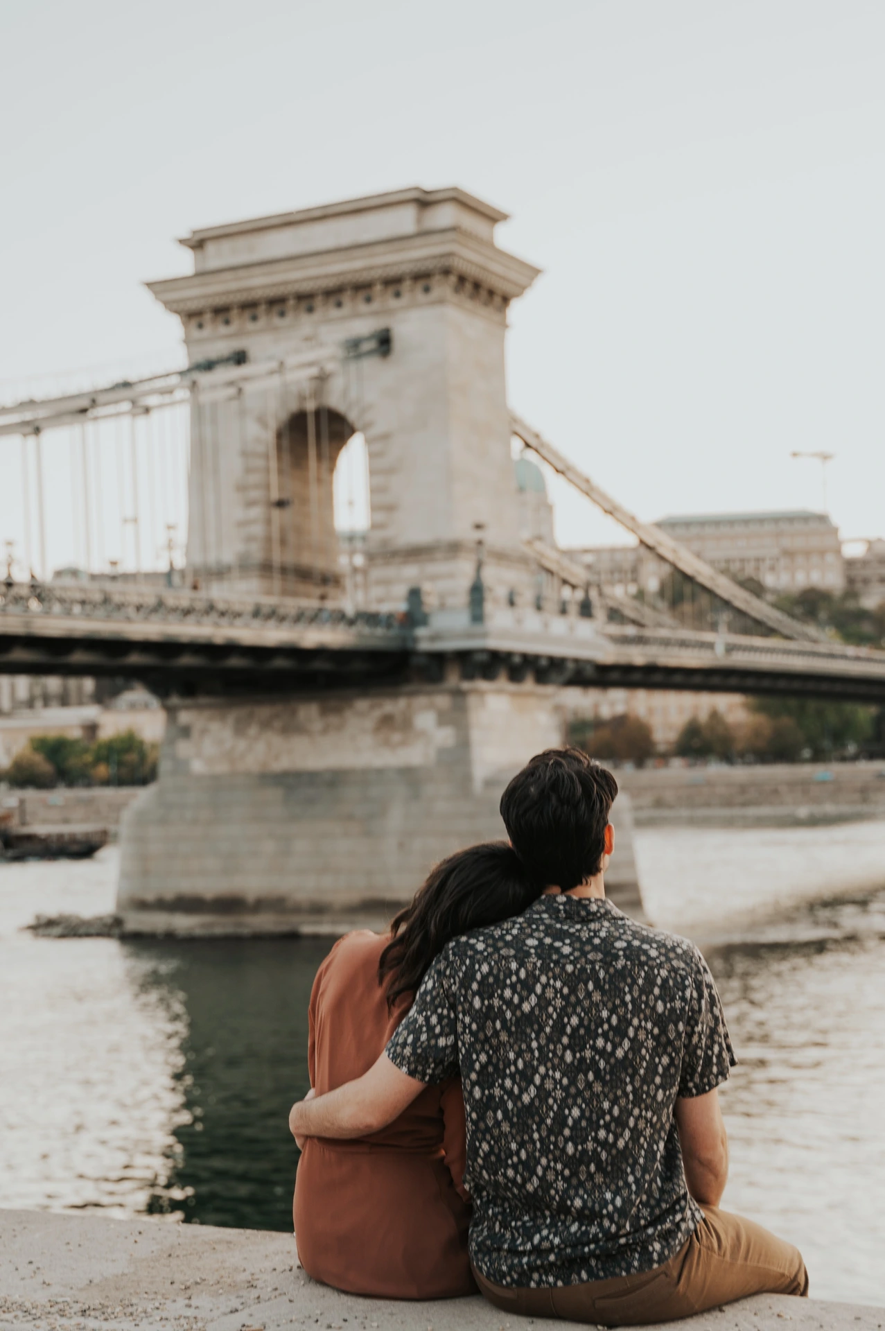 The couple sitting side by side on the riverside stone ledge, seen from behind as they look towards Chain Bridge during their Budapest engagement photoshoot.