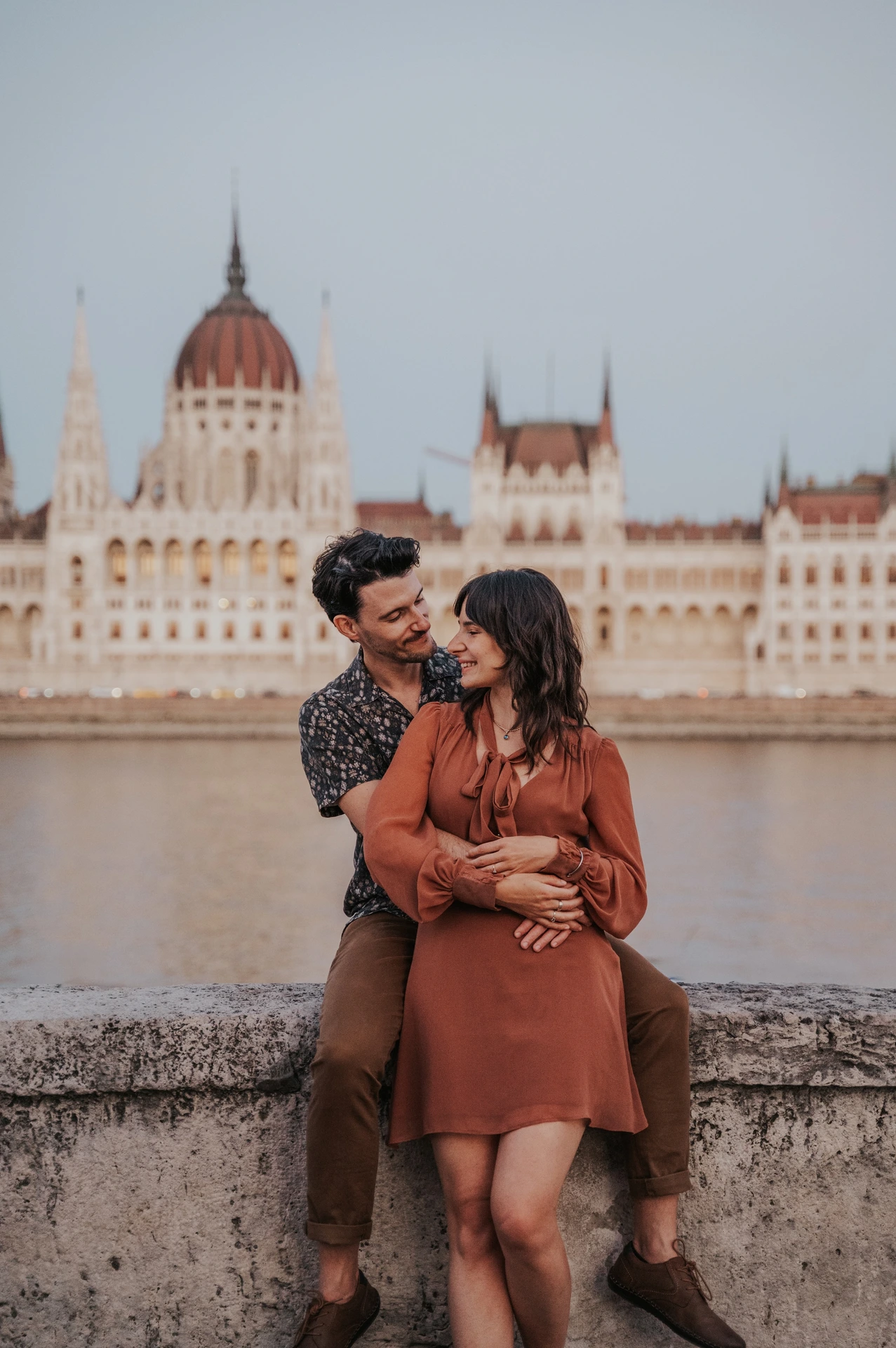 The couple smiling in an embrace beside the riverside wall, with the Hungarian Parliament in the backdrop during their Budapest engagement photoshoot.