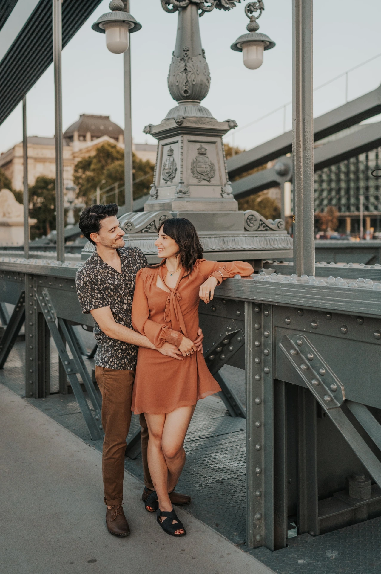 Budapest engagement photoshoot: Standing together beneath a bridge lamp The couple standing close beside the railing under a decorative lamp on Chain Bridge, with its dark metalwork around them during their Budapest engagement photoshoot.