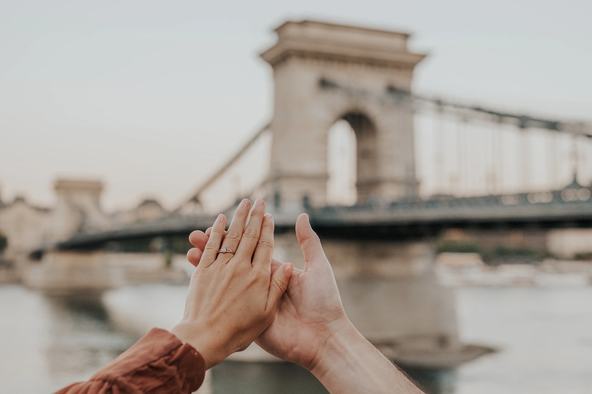 A landscape close-up of the couple’s hands together, showing the engagement ring with Chain Bridge softly blurred across the river behind them during their Budapest engagement photoshoot.