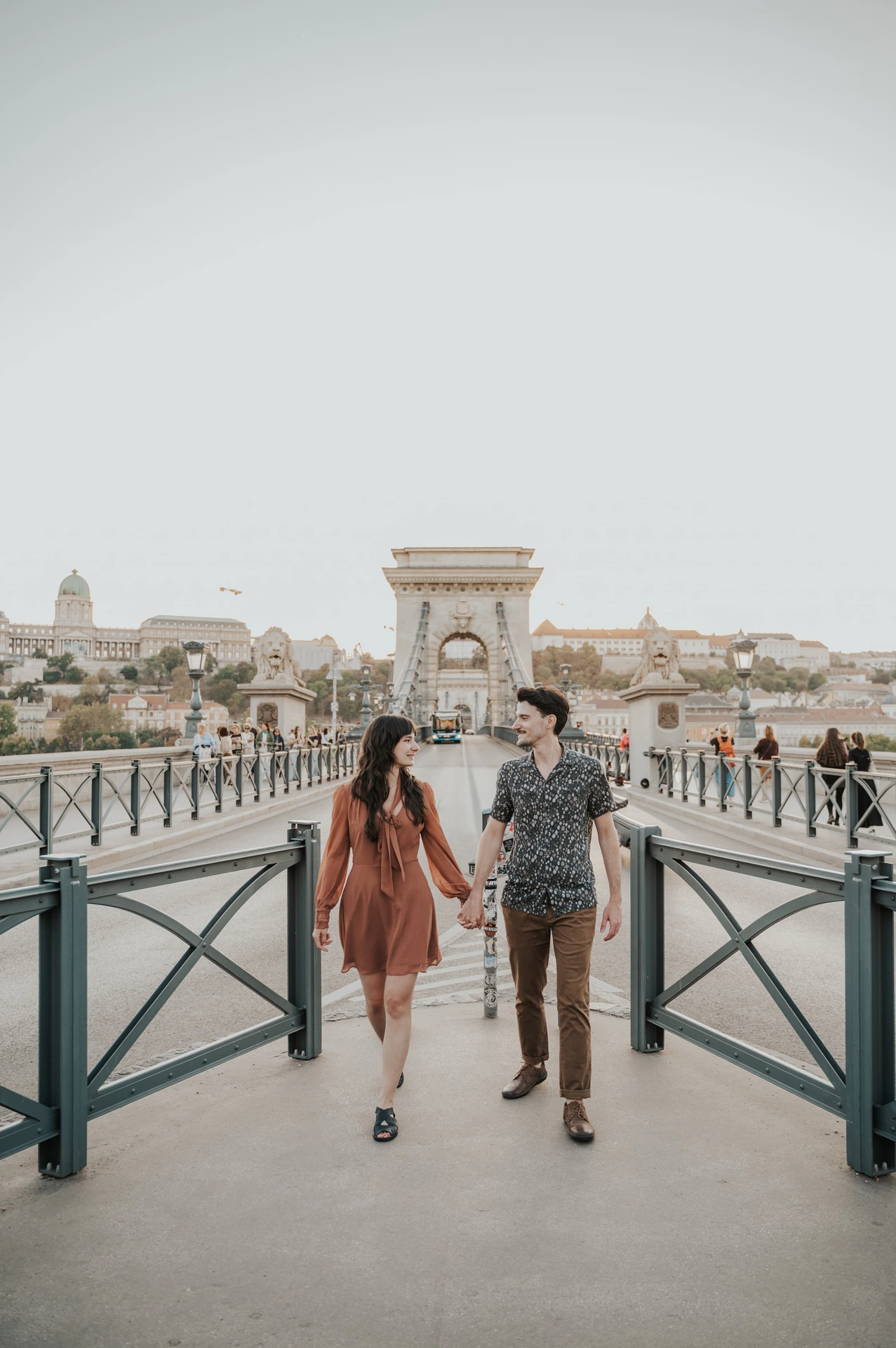 The couple walking hand in hand along Chain Bridge, with its stone tower and dark metal railings behind them during their Budapest engagement photoshoot.