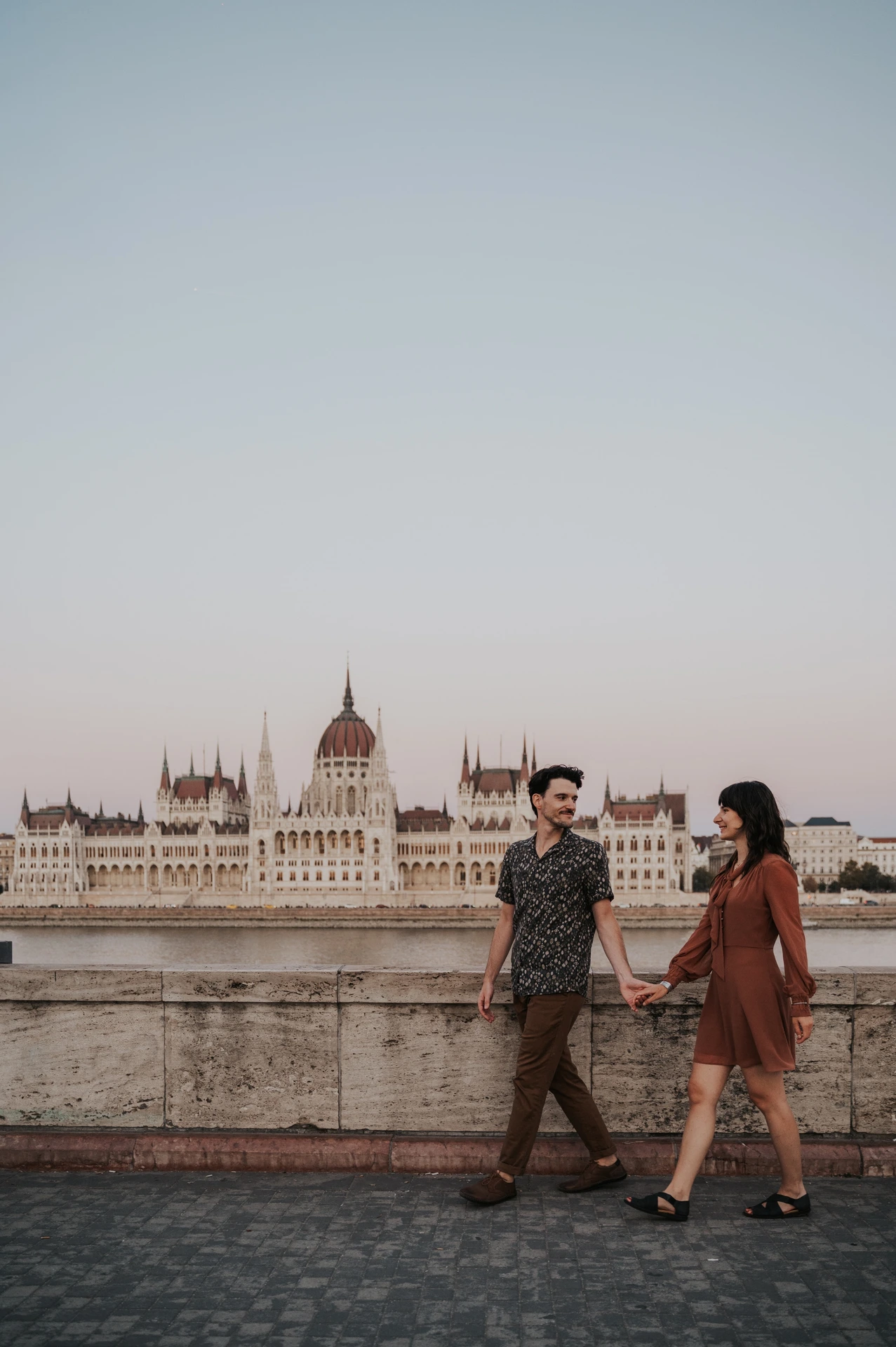 The couple walking hand in hand beside the stone river wall, with the Hungarian Parliament across the Danube during their Budapest engagement photoshoot.