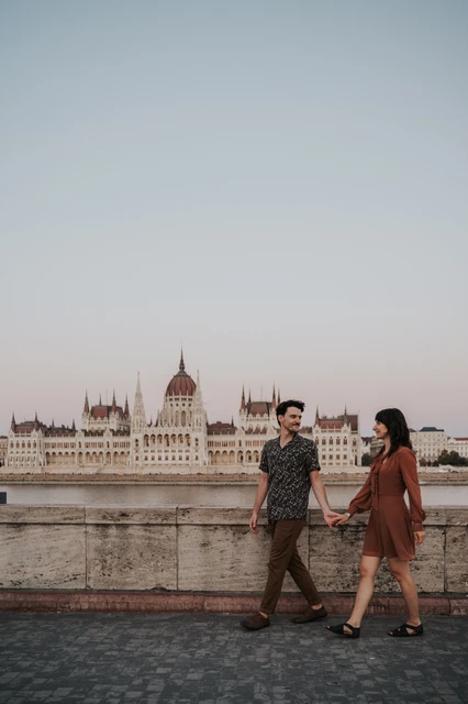Budapest engagement photoshoot: Walking together with Parliament behind them The couple walking hand in hand beside the stone river wall, with the Hungarian Parliament across the Danube during their Budapest engagement photoshoot.