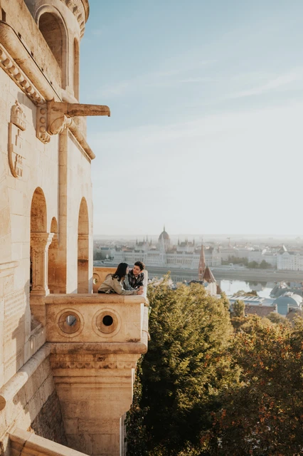 Budapest engagement photoshoot: a balcony portrait with autumn-coloured treetops The couple together on the small Bastion balcony, with sunlit treetops below and Parliament in the distant backdrop during their Budapest engagement photoshoot.