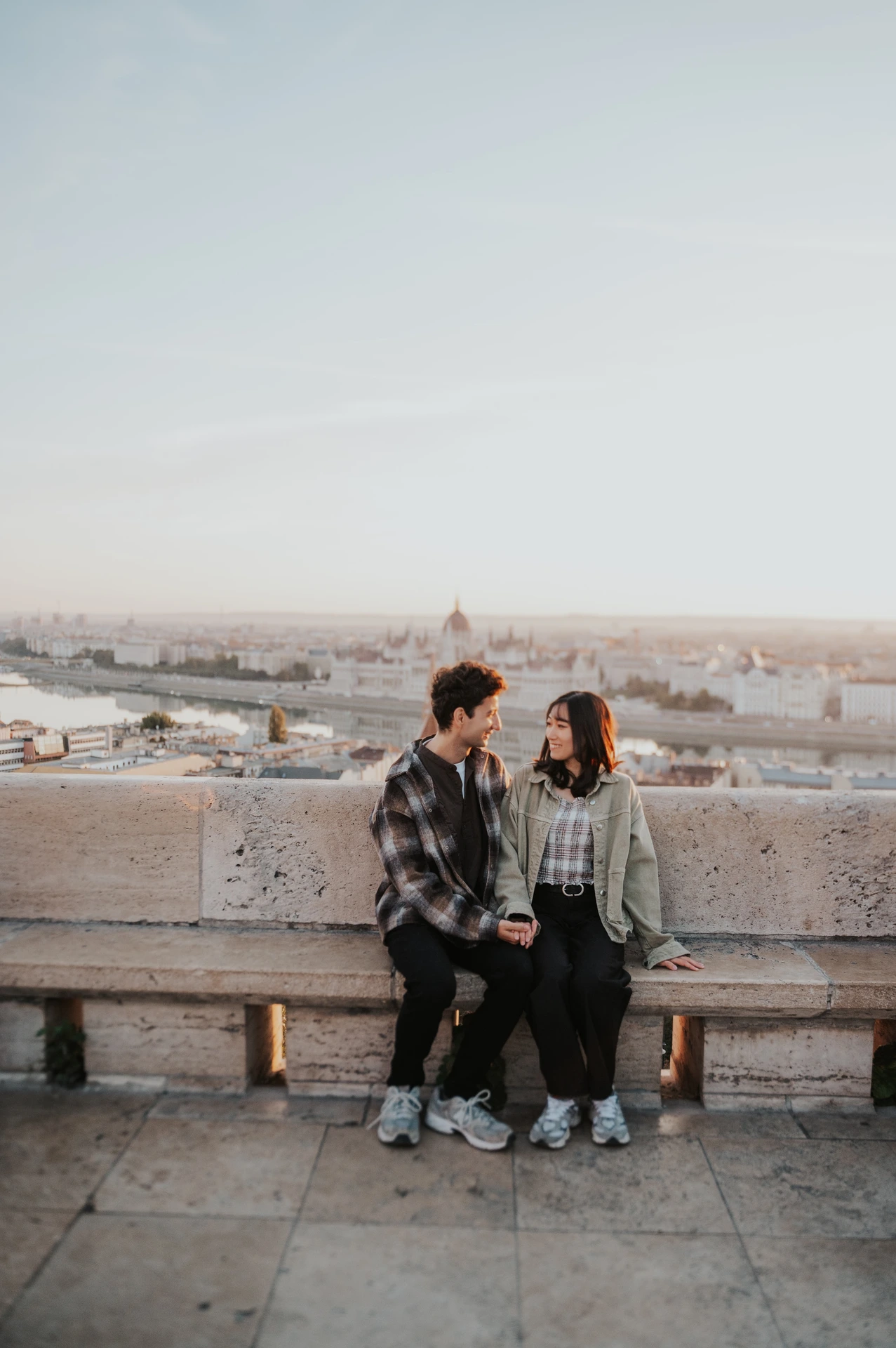 A wide vertical view of the couple seated on a stone bench at Fisherman’s Bastion, with the Budapest skyline stretching behind them during their Budapest engagement photoshoot.