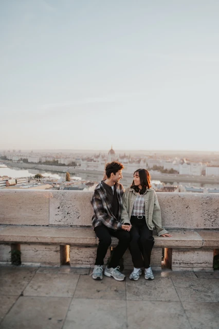 Budapest engagement photoshoot: a bench portrait with Budapest behind them A wide vertical view of the couple seated on a stone bench at Fisherman’s Bastion, with the Budapest skyline stretching behind them during their Budapest engagement photoshoot.