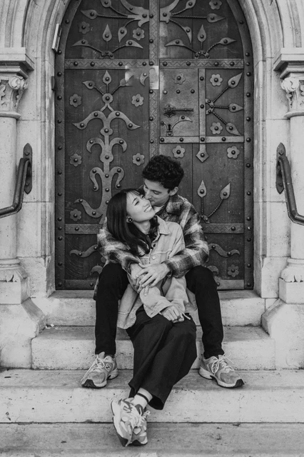Budapest engagement photoshoot: a black-and-white cuddle on the steps Black-and-white portrait of the couple cuddling on the doorway steps, with her leaning back into his arms during their Budapest engagement photoshoot.