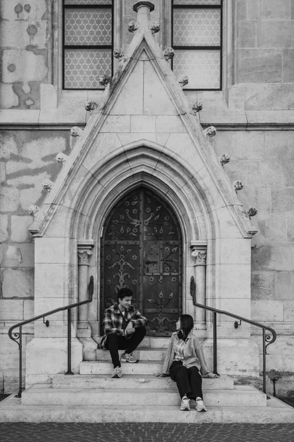 Budapest engagement photoshoot: a black-and-white doorway portrait Black-and-white view of the couple sitting on the stone steps beneath the carved doorway, with curved metal railings beside them during their Budapest engagement photoshoot.