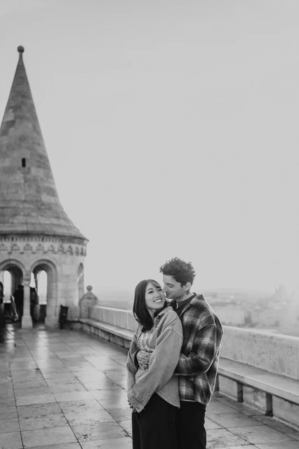 Budapest engagement photoshoot: a black-and-white embrace on the walkway Black-and-white portrait of the couple embracing beside the Bastion’s stone balustrade and pointed tower during their Budapest engagement photoshoot.