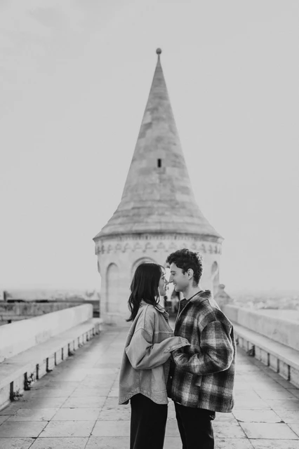 Budapest engagement photoshoot: a black-and-white kiss beside the tower Black-and-white view of the couple sharing a kiss on the Bastion walkway, framed by the tower and stone balustrades during their Budapest engagement photoshoot.