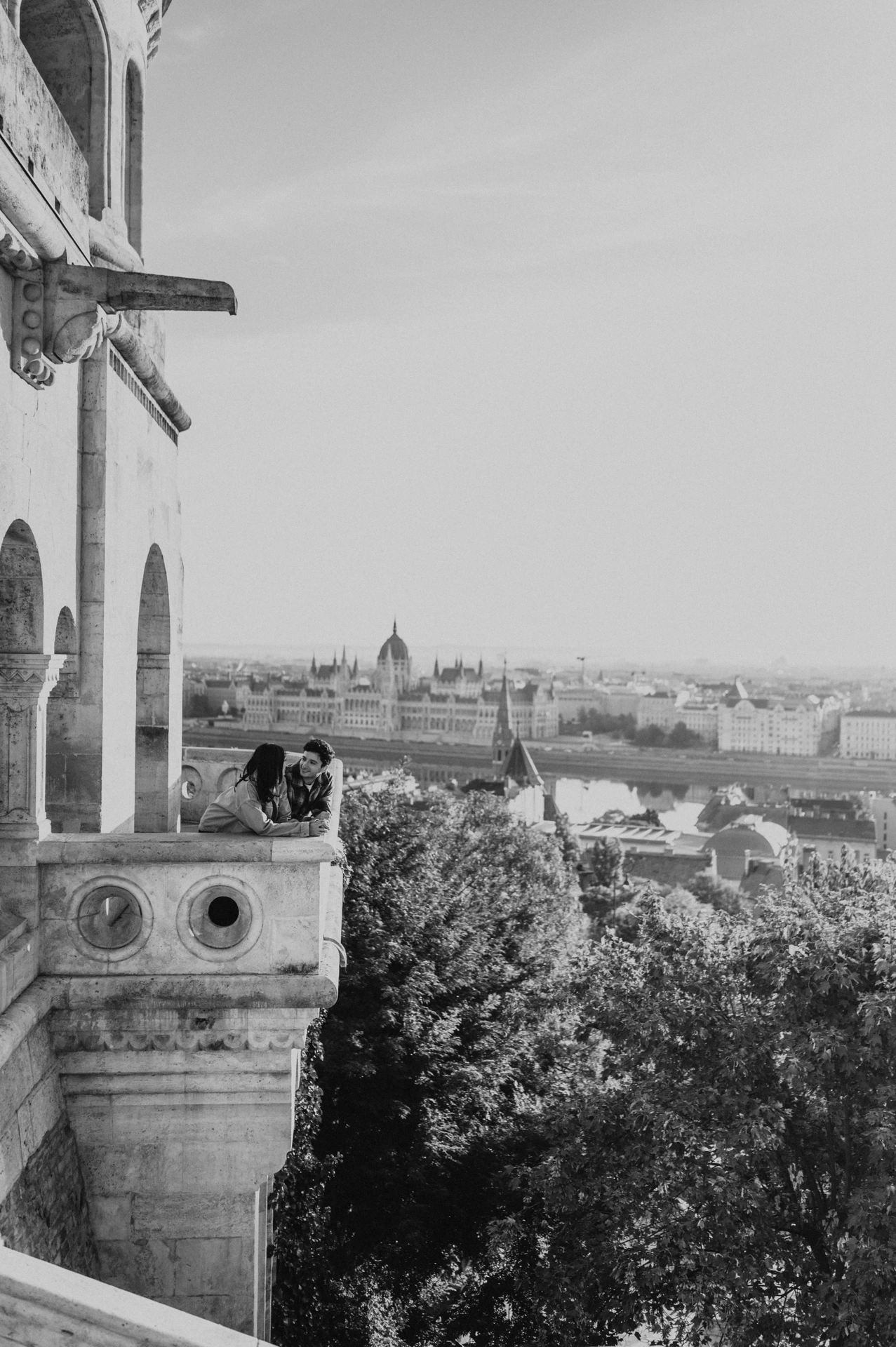 Black-and-white view of the couple kissing at the balcony corner, with Parliament and the river in the distance during their Budapest engagement photoshoot.