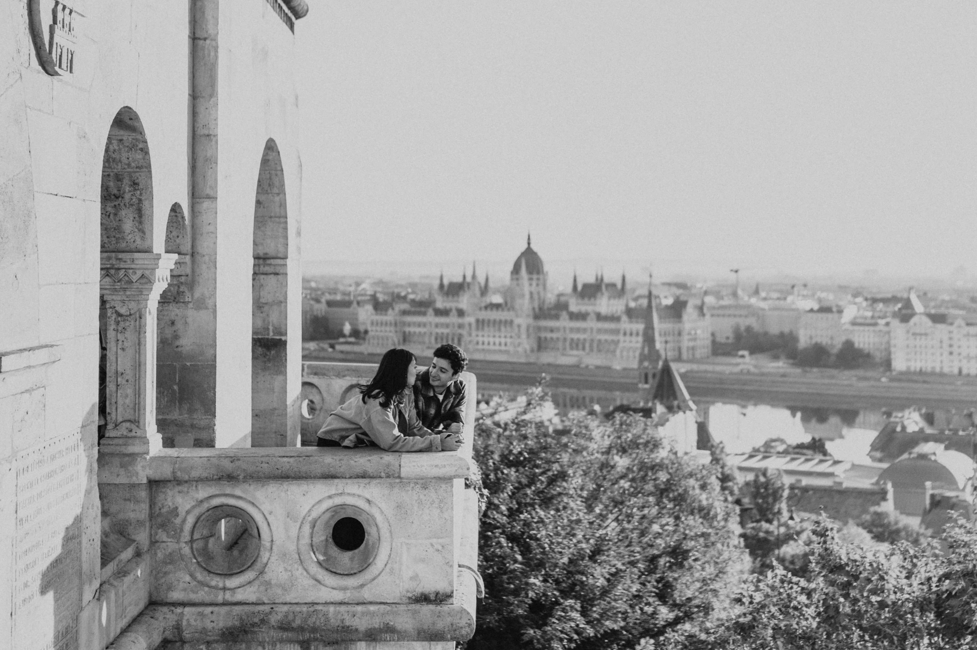 Black-and-white landscape view of the couple kissing at the balcony, with the Danube and Parliament behind them during their Budapest engagement photoshoot.