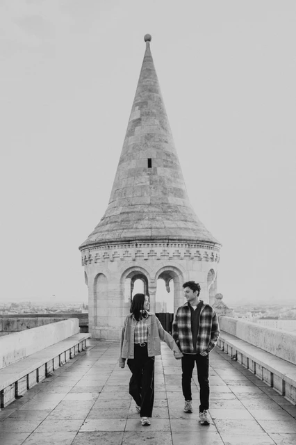Budapest engagement photoshoot: a black-and-white walk towards the camera Black-and-white photograph of the couple walking hand in hand towards the camera on the Bastion walkway during their Budapest engagement photoshoot.