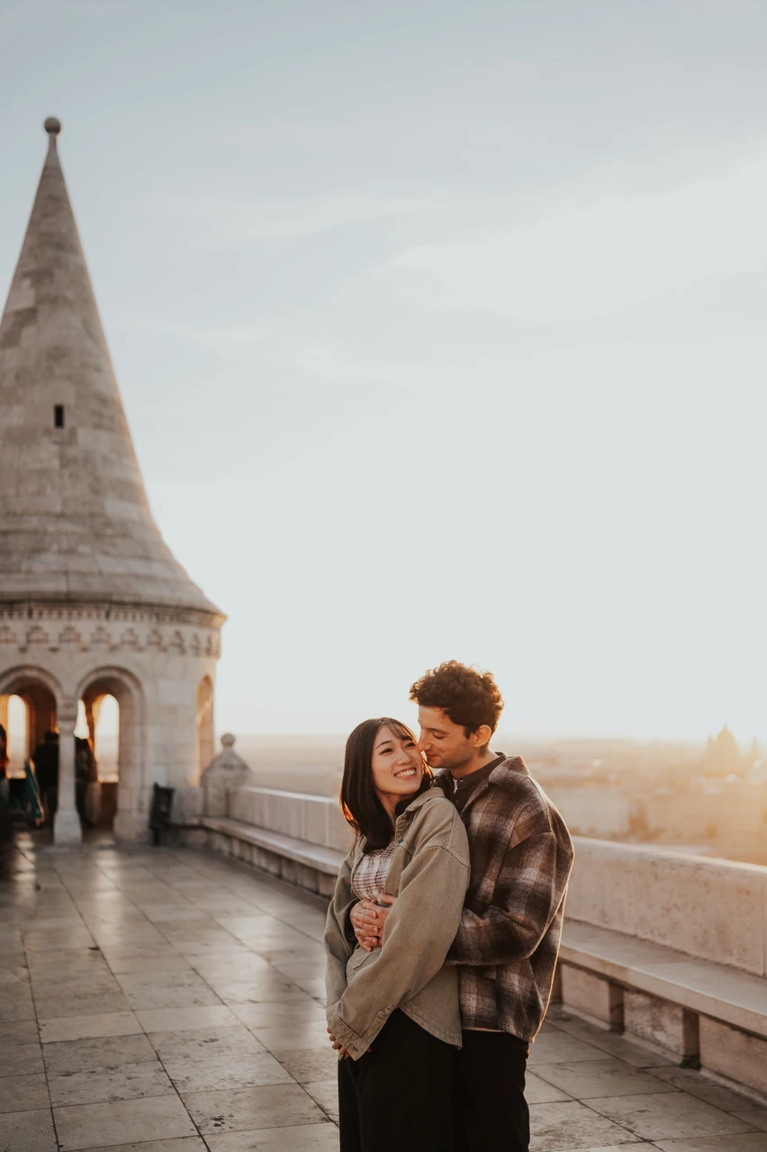 Budapest engagement photoshoot: a cheek kiss in the golden morning light He kisses her cheek while embracing her from behind on the Bastion walkway, with sunlight along the stone railing during their Budapest engagement photoshoot.