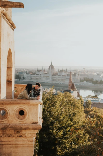 Budapest engagement photoshoot: a close balcony kiss with Parliament behind A close view of the couple kissing over the stone balcony wall, with Parliament and Budapest’s skyline behind them during their Budapest engagement photoshoot.