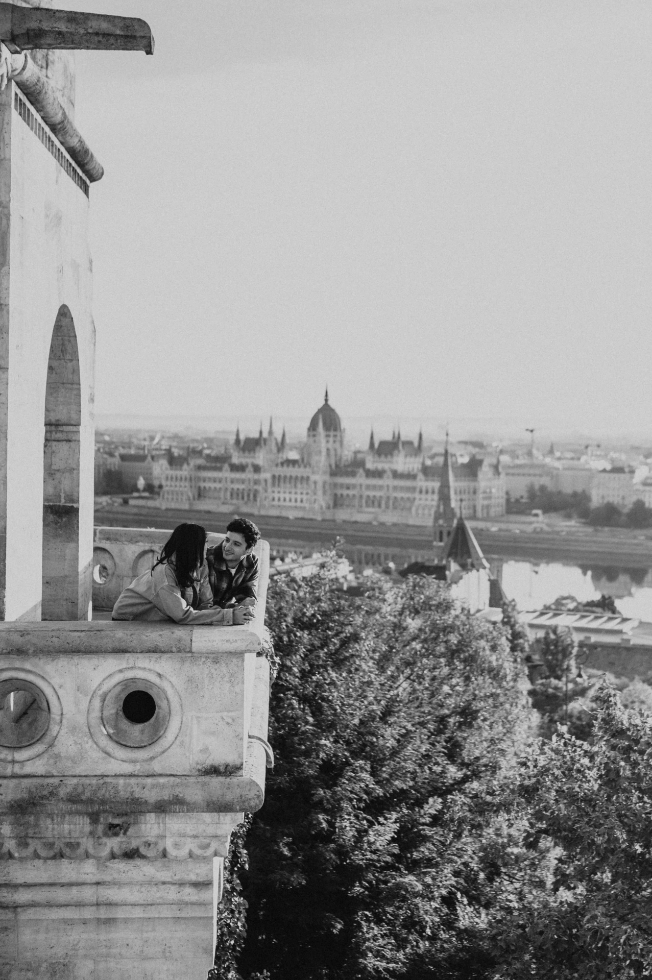 A close black-and-white photograph of the couple kissing on the Bastion balcony, framed by the city and river during their Budapest engagement photoshoot.