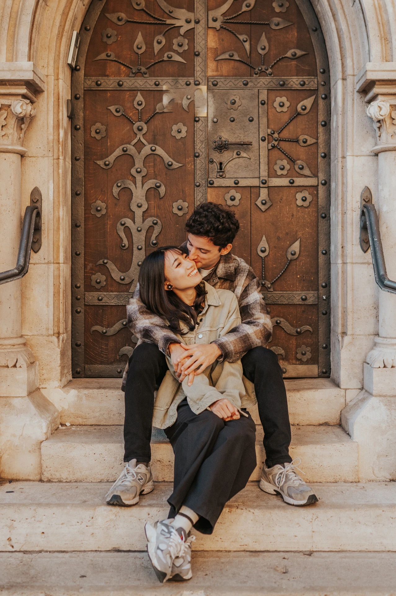 The couple cuddling on the stone steps, with their trainers visible and the decorative wooden door behind them during their Budapest engagement photoshoot.