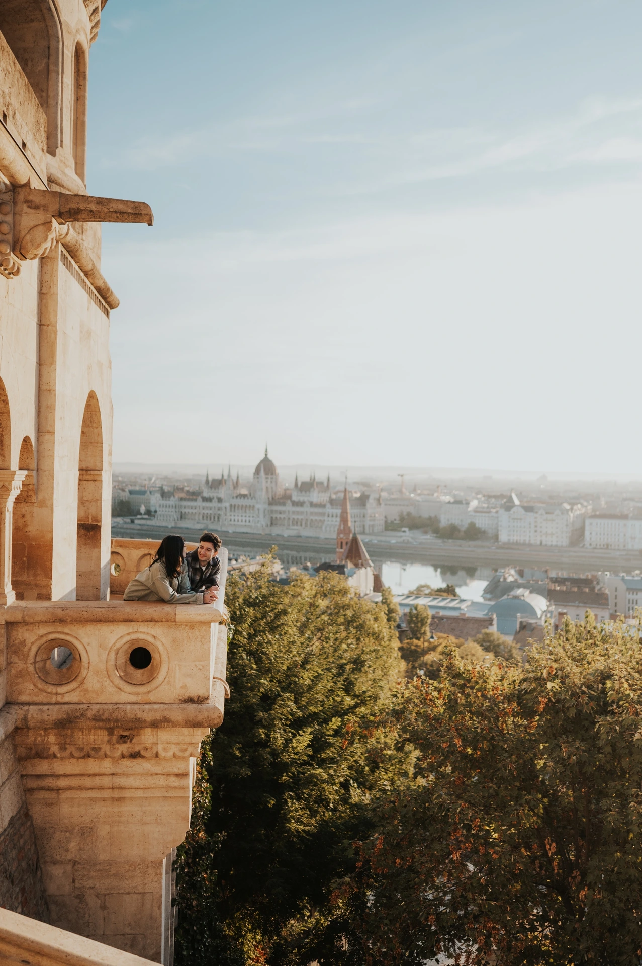 A closer vertical view of the couple beside the stone balcony wall, with Parliament visible across the Danube during their Budapest engagement photoshoot.