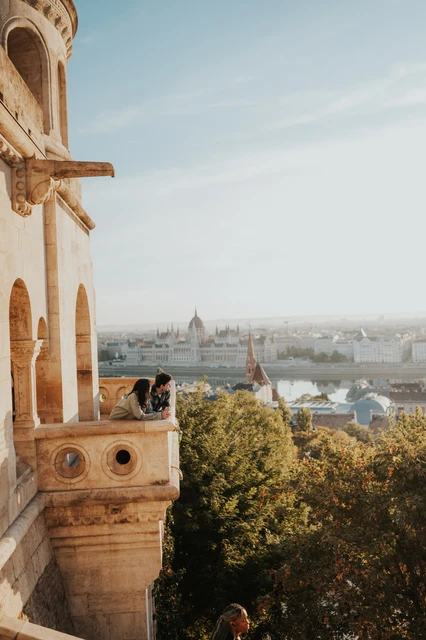 Budapest engagement photoshoot: a full-height view of the balcony and tower The couple leaning together on the balcony at Fisherman’s Bastion, with the full height of the sunlit tower façade above them during their Budapest engagement photoshoot.