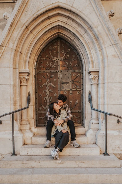 Budapest engagement photoshoot: a full view of the arched doorway and embrace A wide vertical view of the couple embracing on the steps beneath the pointed stone arch and decorative wooden door during their Budapest engagement photoshoot.