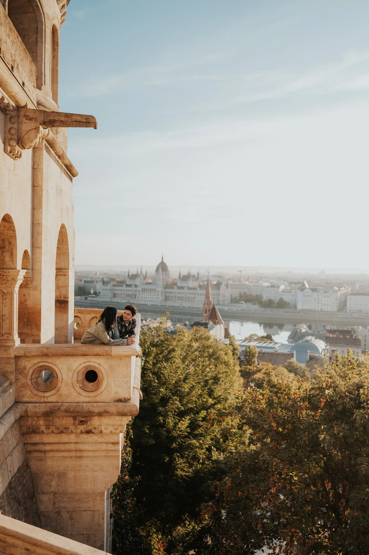 The couple sharing a kiss at the corner of the Bastion balcony, with golden light on the stone and treetops below during their Budapest engagement photoshoot.