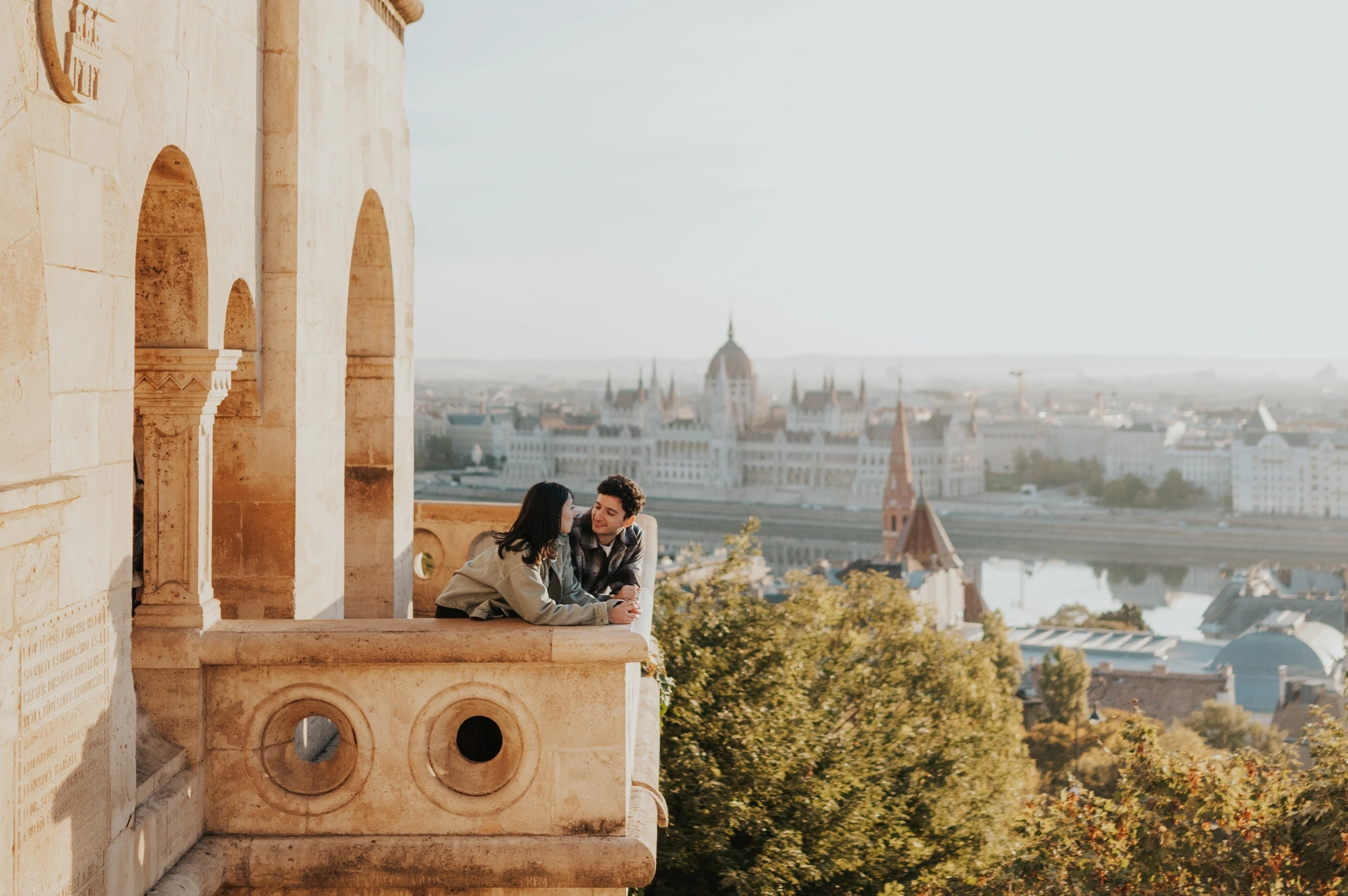 A landscape photograph of the couple kissing on the Bastion balcony, with the stone arch to the left and Parliament across the river during their Budapest engagement photoshoot.