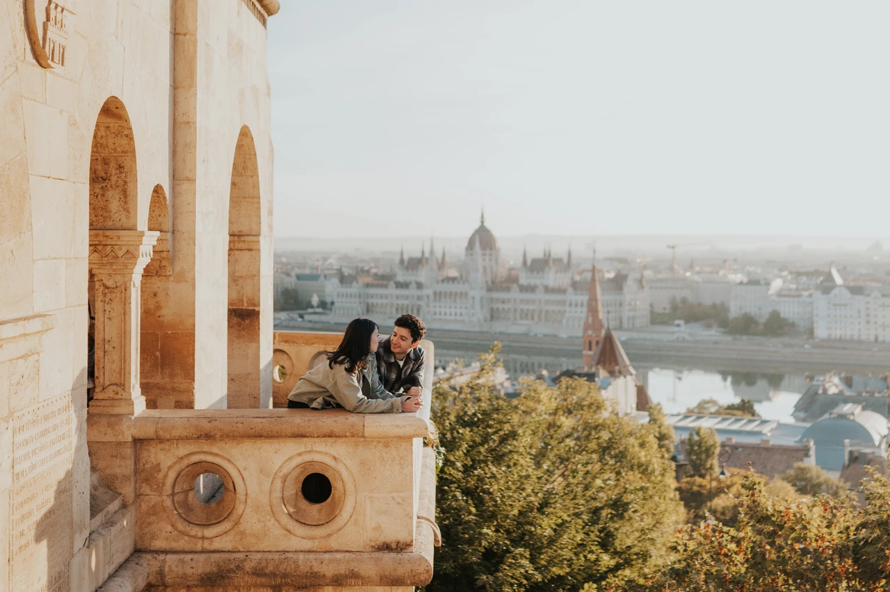 Budapest engagement photoshoot: a landscape balcony kiss with Parliament beyond A landscape photograph of the couple kissing on the Bastion balcony, with the stone arch to the left and Parliament across the river during their Budapest engagement photoshoot.
