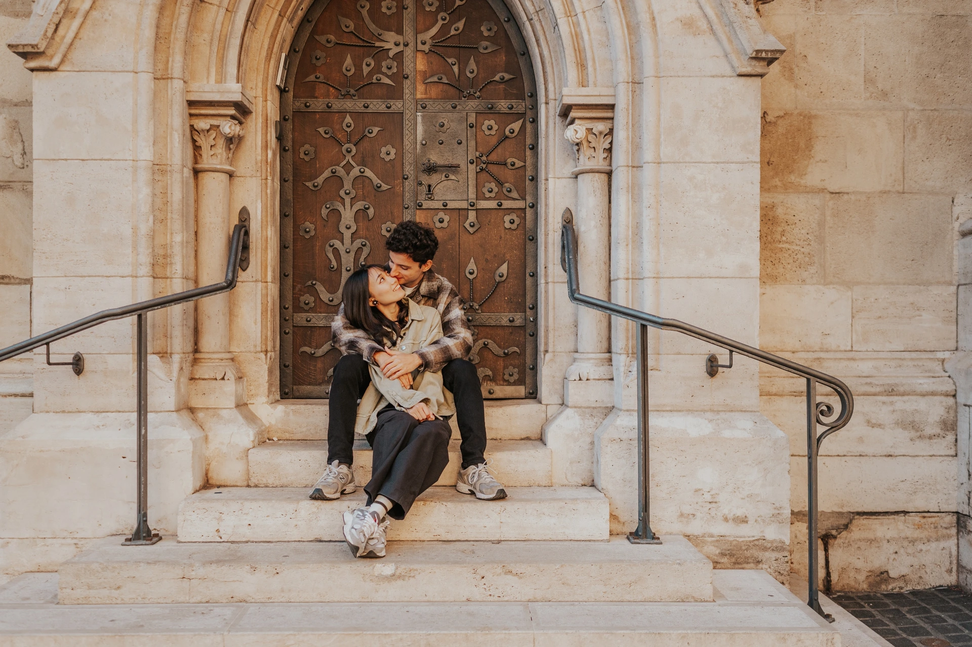 A landscape photograph of the couple cuddling on the stone steps, framed by the ornate doorway and curved metal railings during their Budapest engagement photoshoot.
