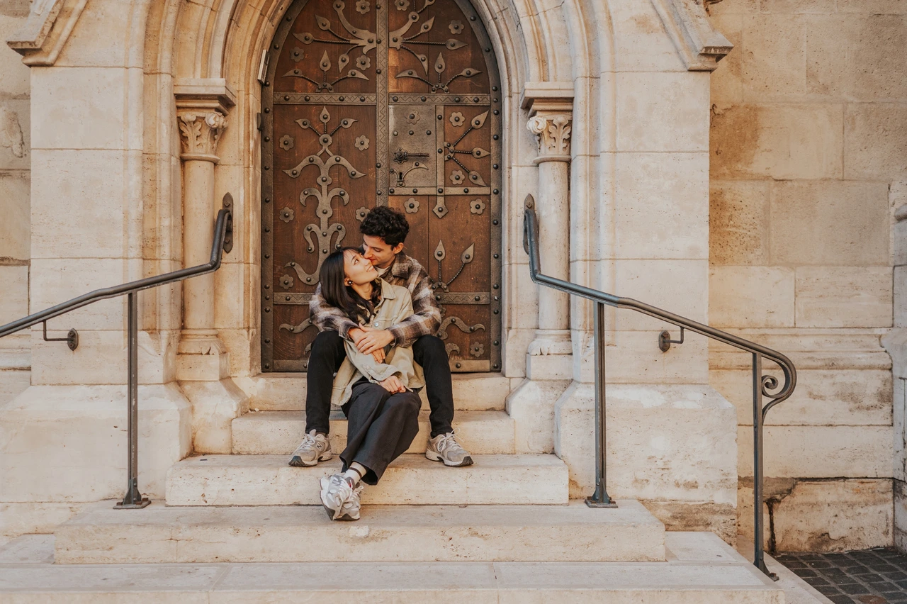 Budapest engagement photoshoot: a landscape cuddle with trainers in view A landscape photograph of the couple cuddling on the stone steps, framed by the ornate doorway and curved metal railings during their Budapest engagement photoshoot.