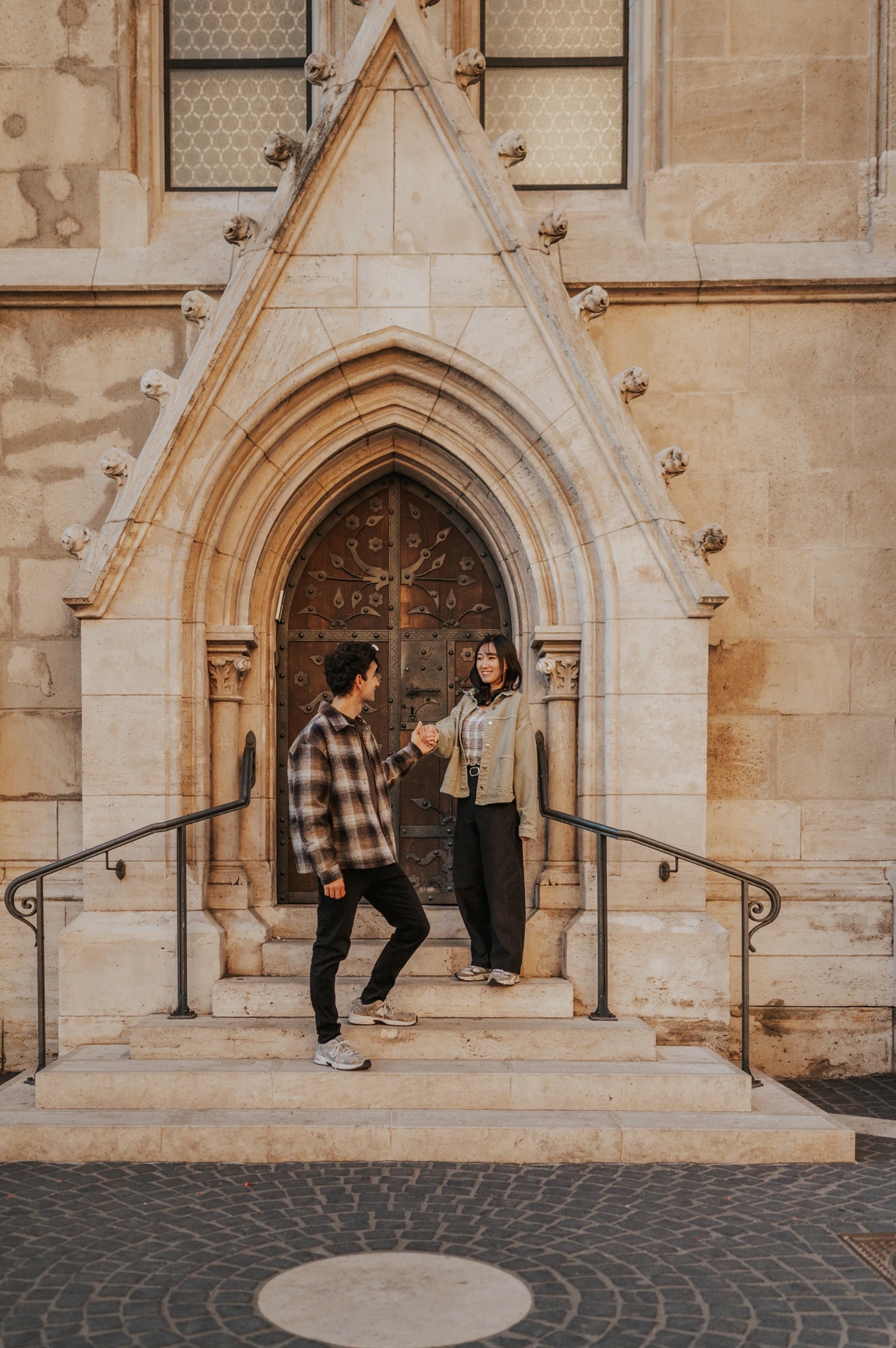 He jumps from the doorway steps while she stands smiling nearby, with the ornate stone arch behind them during their Budapest engagement photoshoot.