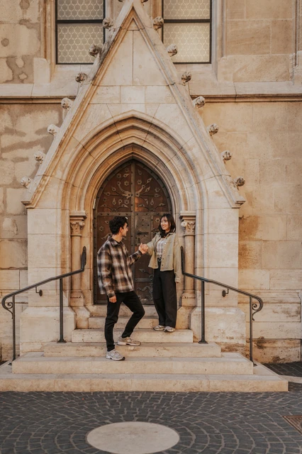 Budapest engagement photoshoot: a playful jump outside the carved doorway He jumps from the doorway steps while she stands smiling nearby, with the ornate stone arch behind them during their Budapest engagement photoshoot.