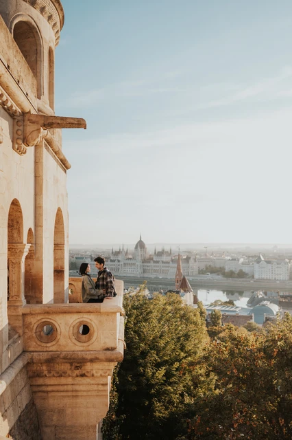 Budapest engagement photoshoot: a seated moment above the treetops The couple seated together on the stone balcony ledge at Fisherman’s Bastion, with leafy treetops and Parliament in the distance during their Budapest engagement photoshoot.