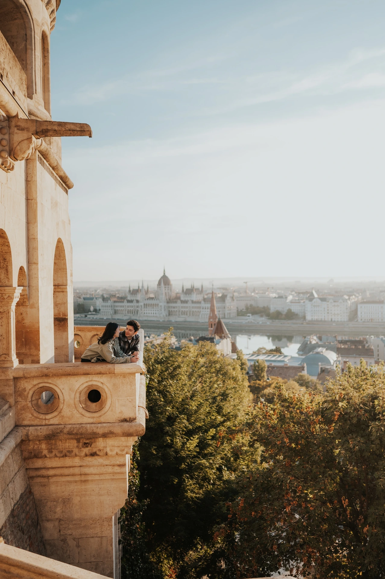The couple facing one another at the curved balcony railing, with warm stonework and the Budapest skyline around them during their Budapest engagement photoshoot.